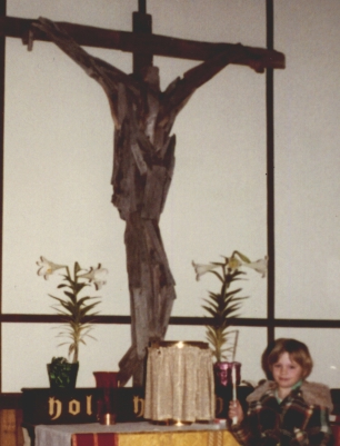 A rough-hewn wooden cross displayed at a church altar, flanked by Easter lilies and candles, a young child sitting nearby